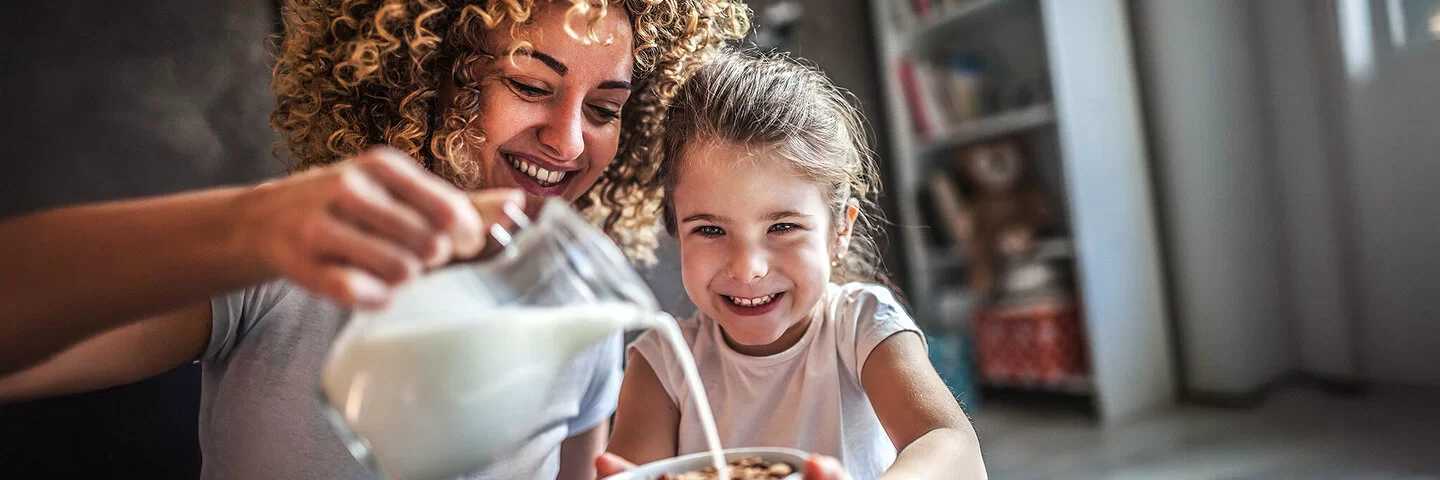 Eine Mutter gießt mit großem Schwung aus einer Karaffe frische Milch in Cornflakes-Schüssel ihrer Tochter.