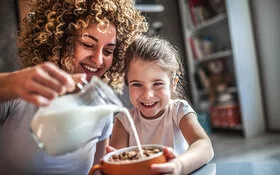 Eine Mutter gießt mit großem Schwung aus einer Karaffe frische Milch in Cornflakes-Schüssel ihrer Tochter.