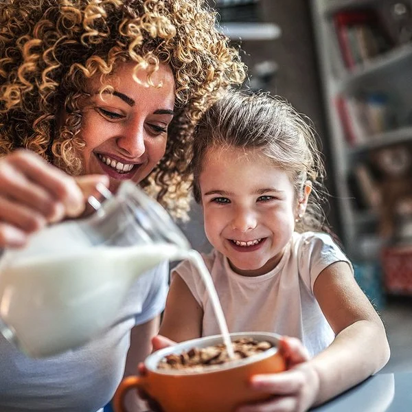 Eine Mutter gießt mit großem Schwung aus einer Karaffe frische Milch in Cornflakes-Schüssel ihrer Tochter.
