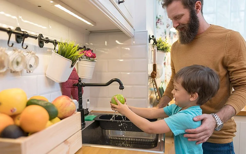 Ein Junge spült einen Apfel unter fließendem Wasser ab, ein Erwachsener steht lächelnd daneben.