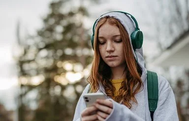 Eine Teenagerin in einem grauen Sweatshirt und grünen Kopfhörern schaut mit ernstem Gesichtsausdruck auf ihr Handy, das sie in den Händen hält.