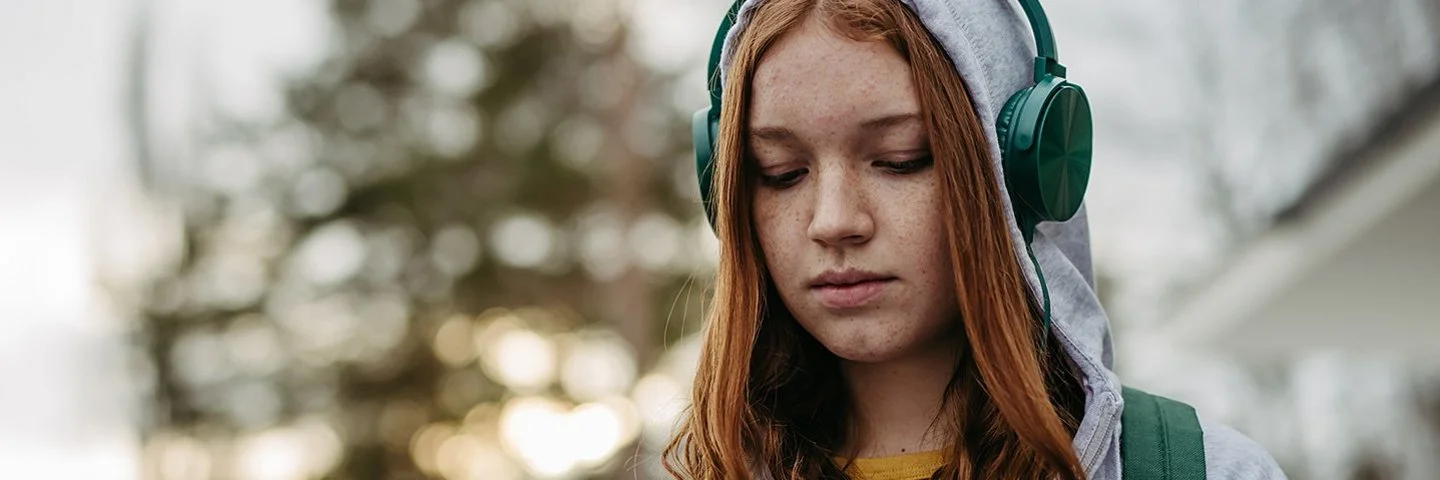 Eine Teenagerin in einem grauen Sweatshirt und grünen Kopfhörern schaut mit ernstem Gesichtsausdruck auf ihr Handy, das sie in den Händen hält.