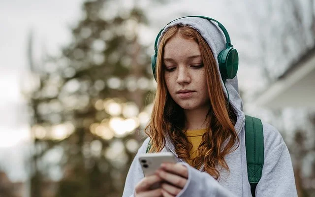 Eine Teenagerin in einem grauen Sweatshirt und grünen Kopfhörern schaut mit ernstem Gesichtsausdruck auf ihr Handy, das sie in den Händen hält.