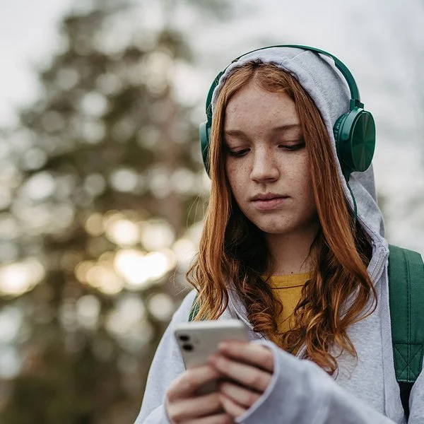 Eine Teenagerin in einem grauen Sweatshirt und grünen Kopfhörern schaut mit ernstem Gesichtsausdruck auf ihr Handy, das sie in den Händen hält.