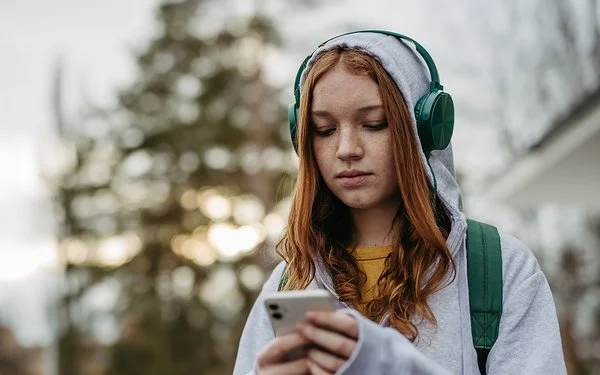 Eine Teenagerin in einem grauen Sweatshirt und grünen Kopfhörern schaut mit ernstem Gesichtsausdruck auf ihr Handy, das sie in den Händen hält.