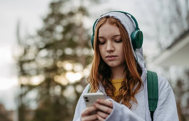 Eine Teenagerin in einem grauen Sweatshirt und grünen Kopfhörern schaut mit ernstem Gesichtsausdruck auf ihr Handy, das sie in den Händen hält.