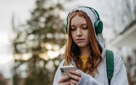 Eine Teenagerin in einem grauen Sweatshirt und grünen Kopfhörern schaut mit ernstem Gesichtsausdruck auf ihr Handy, das sie in den Händen hält.