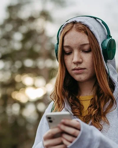 Eine Teenagerin in einem grauen Sweatshirt und grünen Kopfhörern schaut mit ernstem Gesichtsausdruck auf ihr Handy, das sie in den Händen hält.