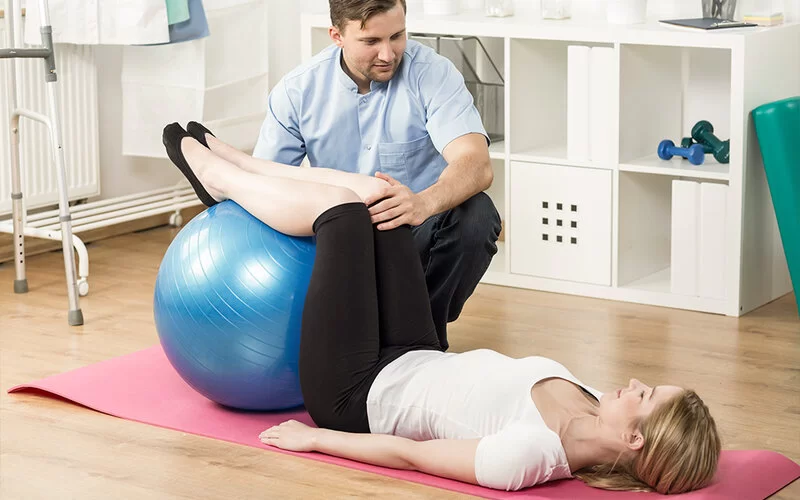 Ein Physiotherapeut zeigt einer Patientin mit Hexenschuss die Stufenlage auf einem Gymnastikball. Die Frau liegt dabei auf einer Sportmatte mit den Beinen auf einem Gymnastikball.