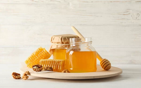 Auf einem Tisch steht ein Glas Honig umgeben von einigen Honigwaben und Walnüssen sowie einem Honiglöffel.