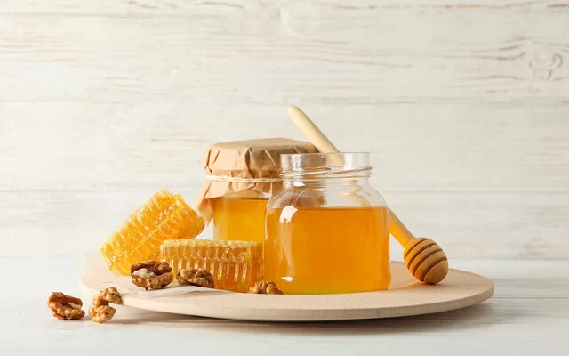 Auf einem Tisch steht ein Glas Honig umgeben von einigen Honigwaben und Walnüssen sowie einem Honiglöffel.