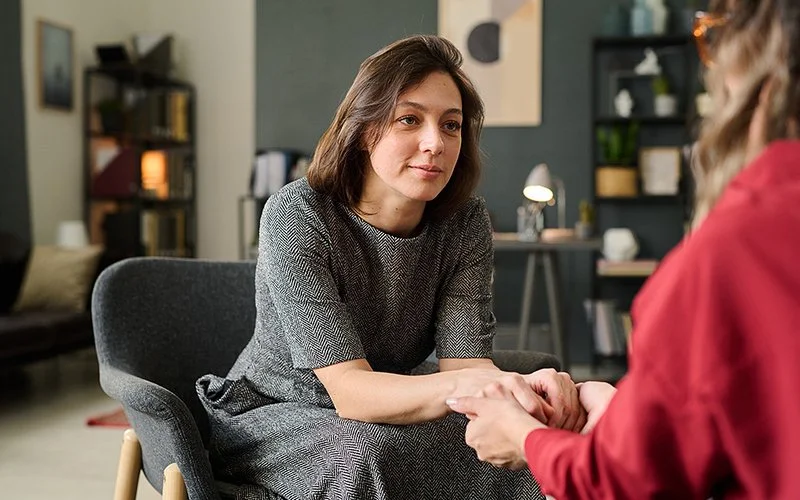 In einem Raum sitzen sich zwei Frauen gegenüber. Eine der Frauen ist nur von hinten im Ausschnitt abgebildet. Sie halten sich an den Händen und schauen sich in die Augen.
