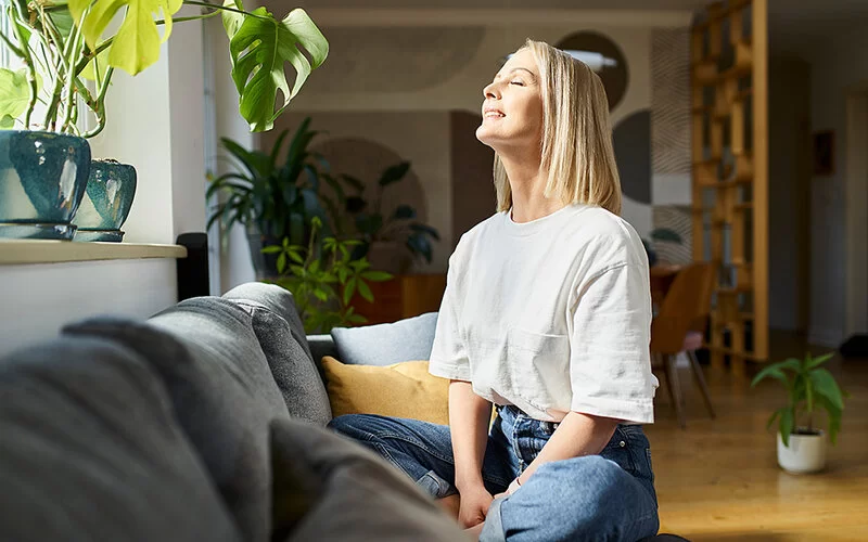 Gegen die Reiseangst: Eine Frau sitzt im Schneidersitz auf dem Sofa macht eine mentale Entspannungsübung.