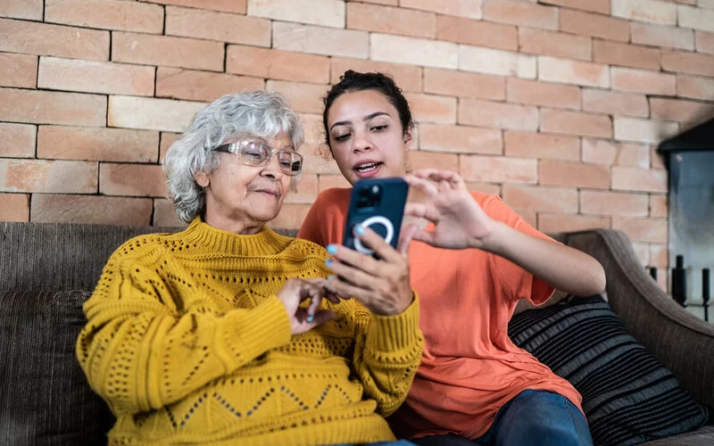 Eine junge Frau sitzt neben einer Seniorin auf einem Sofa und hilft ihr, mit einem Smartphone umzugehen.