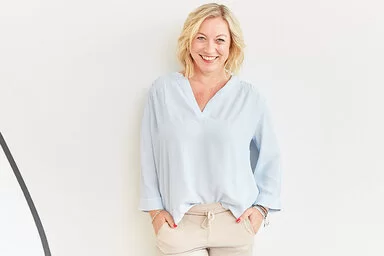 Coach Angela Homfeldt mit schulterlangen blonden Haaren steht in einer hellblauen Bluse und einer beigefarbenen Hose vor einer weißen Wand und lächelt in die Kamera. 