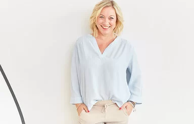 Coach Angela Homfeldt mit schulterlangen blonden Haaren steht in einer hellblauen Bluse und einer beigefarbenen Hose vor einer weißen Wand und lächelt in die Kamera. 