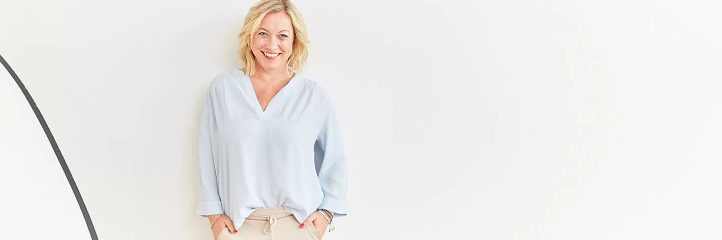 Coach Angela Homfeldt mit schulterlangen blonden Haaren steht in einer hellblauen Bluse und einer beigefarbenen Hose vor einer weißen Wand und lächelt in die Kamera.
