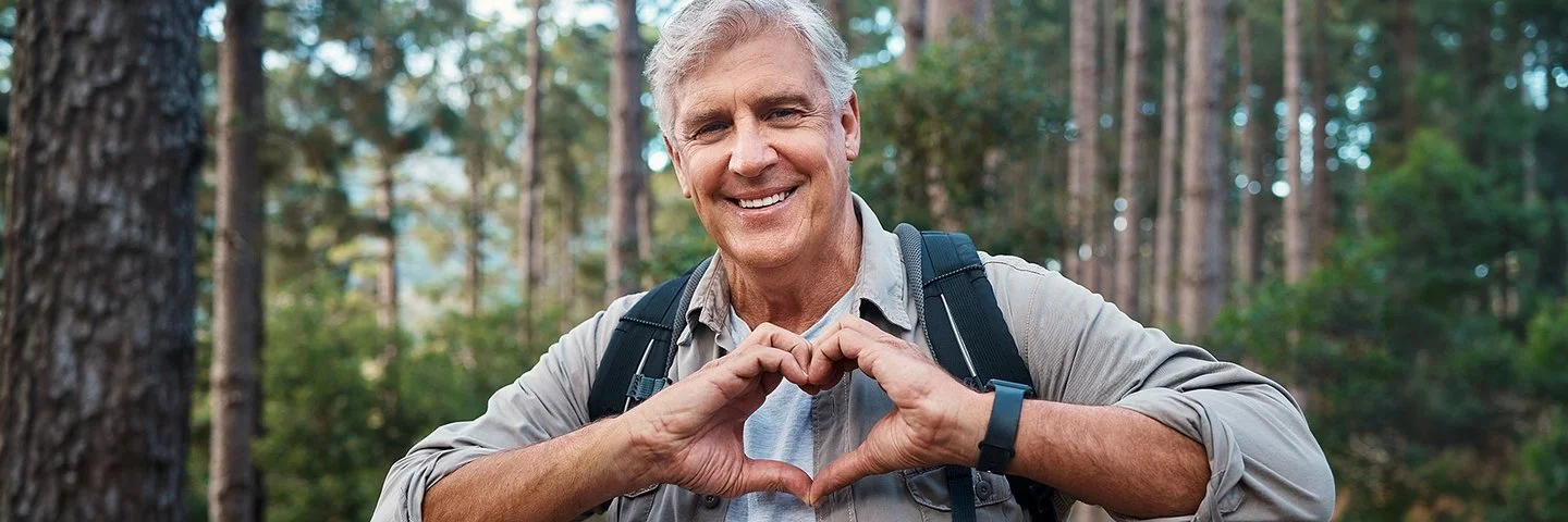 Ein älterer Mann ist im Wald unterwegs und formt seine Hände zu einem Herz.