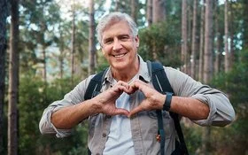 Ein älterer Mann ist im Wald unterwegs und formt seine Hände zu einem Herz.