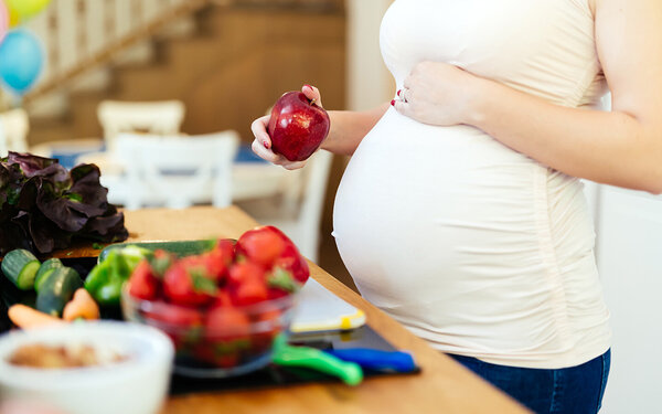 Eine schwangere Frau hält einen Apfel in der Hand und ist dabei, einen Salat zuzubereiten.