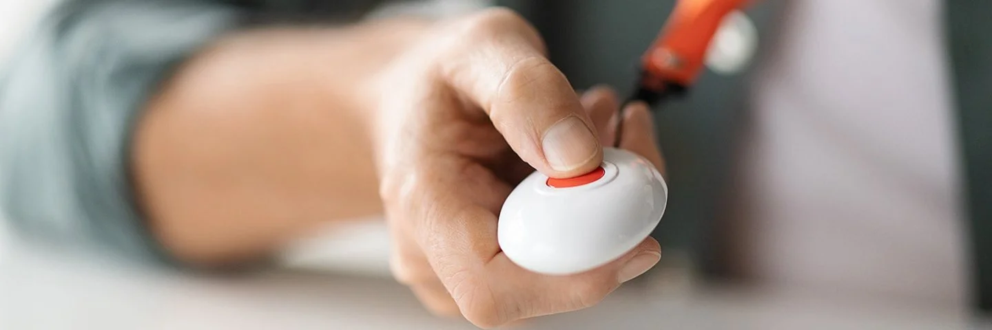 Eine Person hält einen kleinen weißen Notfallknopf mit roter Taste in der Hand, der an einem orangefarbenen Band um den Hals getragen wird.