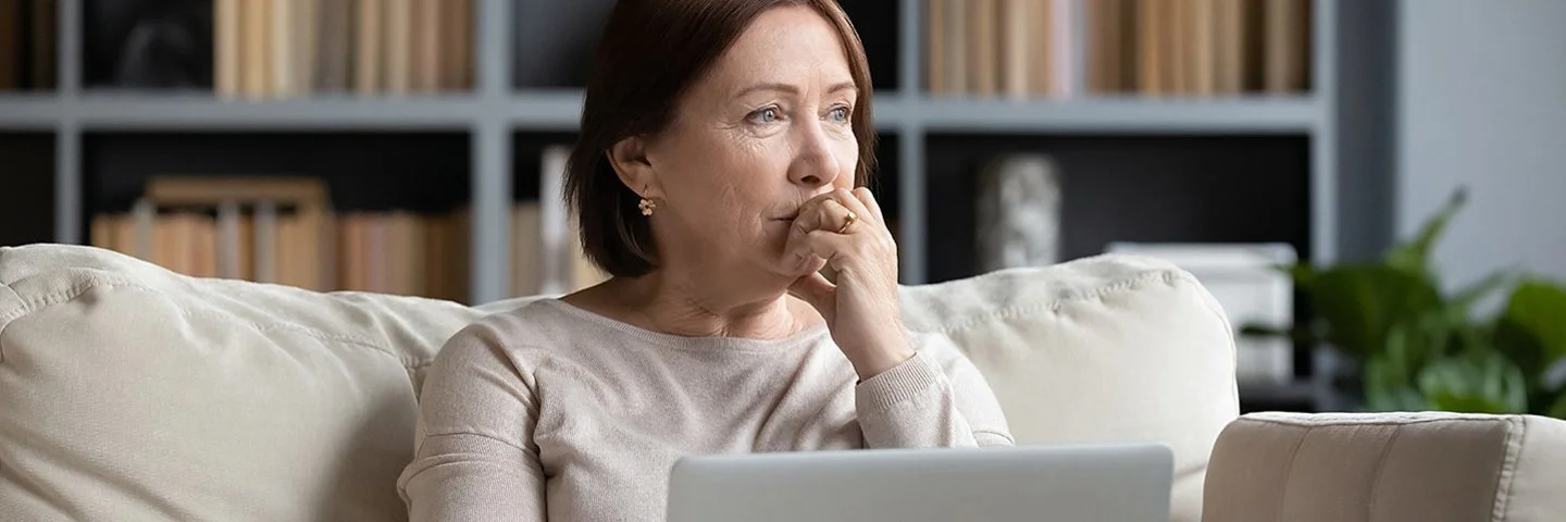 Ältere Frau sitzt mit dem Laptop auf dem Schoß auf einer Couch und schaut nachdenklich ins Leere.
