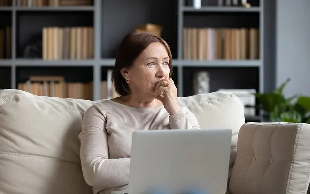 Ältere Frau sitzt mit dem Laptop auf dem Schoß auf einer Couch und schaut nachdenklich ins Leere.