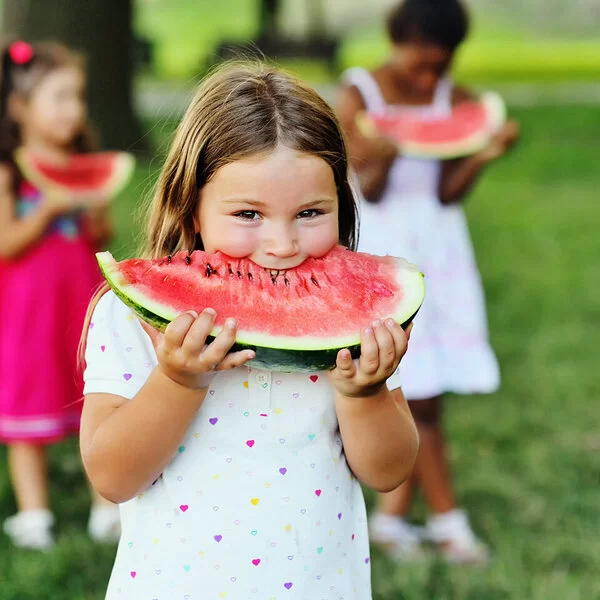 Ein kleines Mädchen steht in einem weißen Sommerkleid mit bunten Herzchen auf einer Wiese in einem Park und beißt genießerisch in eine Wassermelonenspalte. Hinter ihr sind zwei weitere Mädchen in bunten Kleidern zu sehen.