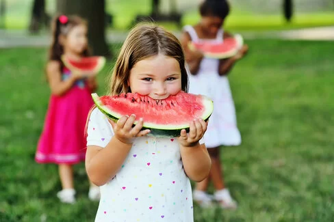 Ein kleines Mädchen steht in einem weißen Sommerkleid mit bunten Herzchen auf einer Wiese in einem Park und beißt genießerisch in eine Wassermelonenspalte. Hinter ihr sind zwei weitere Mädchen in bunten Kleidern zu sehen.