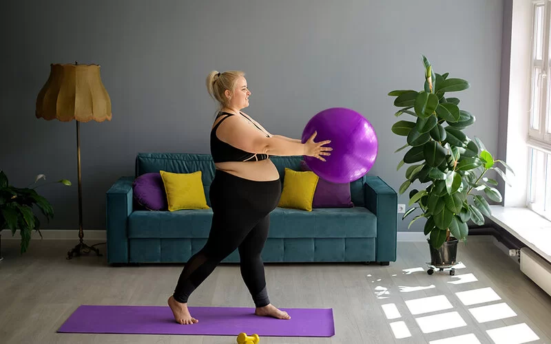Eine von Übergewicht betroffene Schwangere in Sportkleidung steht auf einer Gymnastikmatte und hält einen Gymnastikball in beiden Händen.
