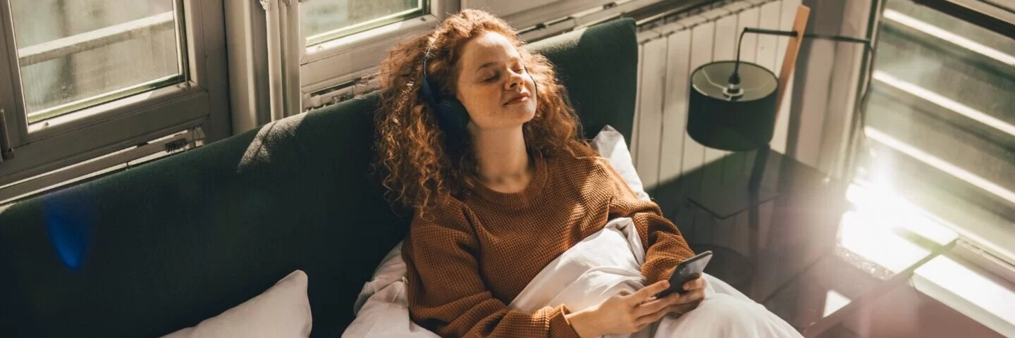 Eine junge Frau sitzt im Bett und trägt Kopfhörer, über die sie ASMR-Geräusche hört.