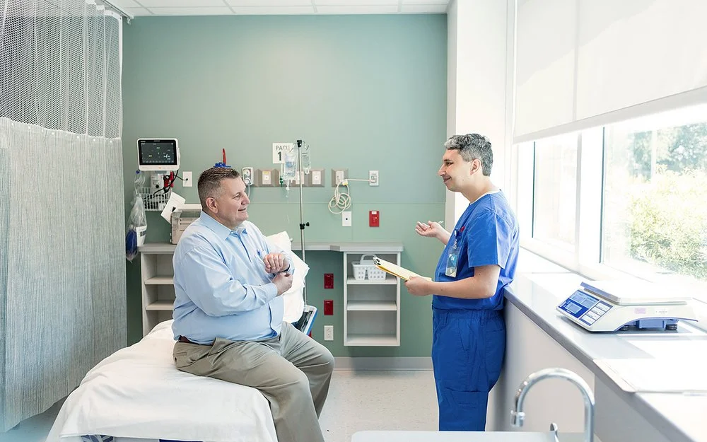 Ein von Übergewicht betroffener Mann sitzt auf einer Liege in einem Untersuchungsraum einer Klinik oder eines medizinischen Zentrums. Er unterhält sich mit einem Arzt, der ihm gegenübersteht.