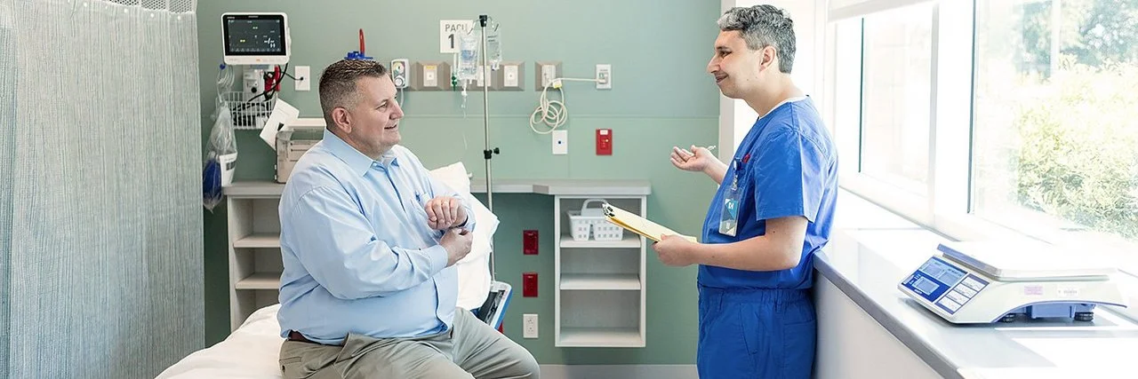 Ein von Übergewicht betroffener Mann sitzt auf einer Liege in einem Untersuchungsraum einer Klinik oder eines medizinischen Zentrums. Er unterhält sich mit einem Arzt, der ihm gegenübersteht.