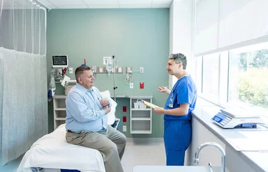 Ein von Übergewicht betroffener Mann sitzt auf einer Liege in einem Untersuchungsraum einer Klinik oder eines medizinischen Zentrums. Er unterhält sich mit einem Arzt, der ihm gegenübersteht.