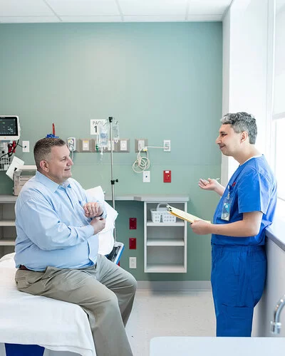 Ein von Übergewicht betroffener Mann sitzt auf einer Liege in einem Untersuchungsraum einer Klinik oder eines medizinischen Zentrums. Er unterhält sich mit einem Arzt, der ihm gegenübersteht.