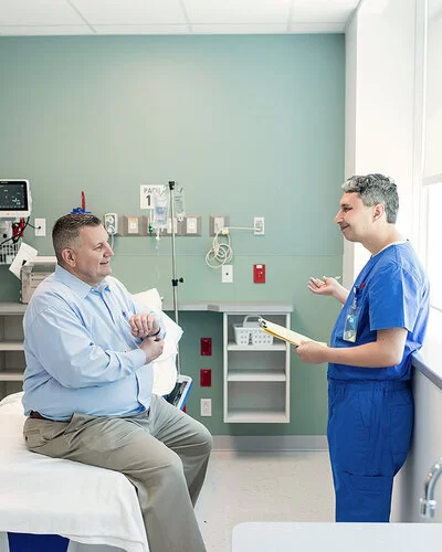 Ein von Übergewicht betroffener Mann sitzt auf einer Liege in einem Untersuchungsraum einer Klinik oder eines medizinischen Zentrums. Er unterhält sich mit einem Arzt, der ihm gegenübersteht.