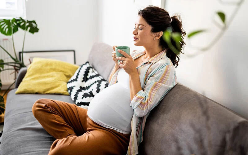 Eine hochschwangere junge Frau sitzt im Schneidersitz auf der Couch und trinkt mit Genuss und geschlossenen Augen einen Tee zur Entspannung.