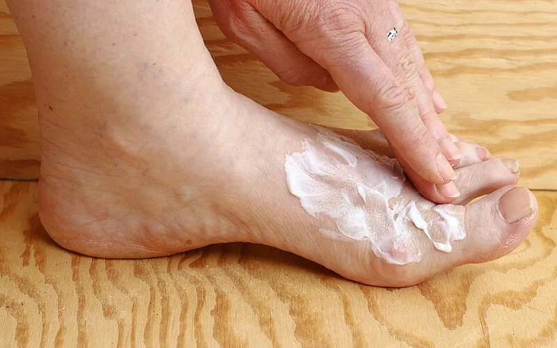 Eine Frau reibt sich den Fuß mit einer weißen Salbe oder Creme ein.