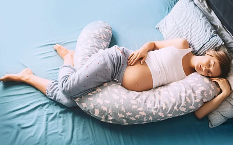 Eine schwangere Frau liegt im Bett auf Seite mit einem Kissen zwischen den Beinen, um Rückenschmerzen zu reduzieren.