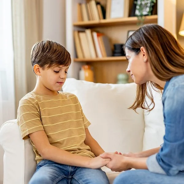 Ein Junge sitzt auf einem weißen Sofa und schaut etwas bedrückt nach unten. Eine Frau lehnt sich zu ihm und hält einfühlsam seine Hände.