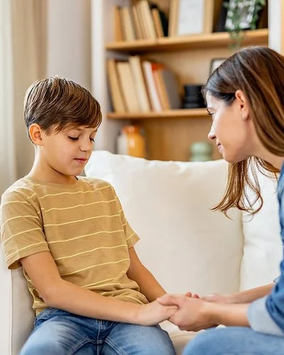 Ein Junge sitzt auf einem weißen Sofa und schaut etwas bedrückt nach unten. Eine Frau lehnt sich zu ihm und hält einfühlsam seine Hände.