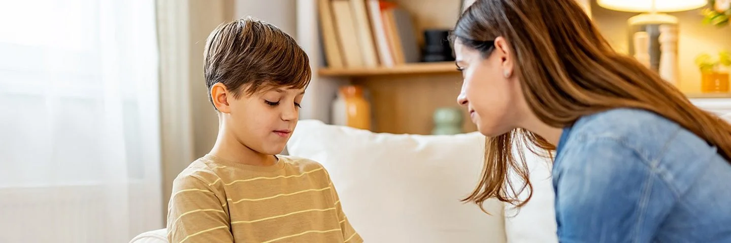 Ein Junge sitzt auf einem weißen Sofa und schaut etwas bedrückt nach unten. Eine Frau lehnt sich zu ihm und hält einfühlsam seine Hände.
