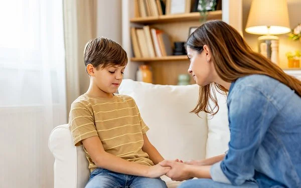 Ein Junge sitzt auf einem weißen Sofa und schaut etwas bedrückt nach unten. Eine Frau lehnt sich zu ihm und hält einfühlsam seine Hände.