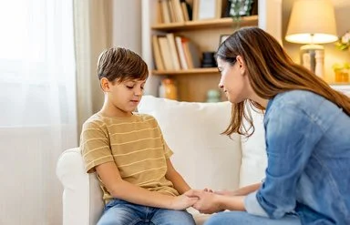Ein Junge sitzt auf einem weißen Sofa und schaut etwas bedrückt nach unten. Eine Frau lehnt sich zu ihm und hält einfühlsam seine Hände.