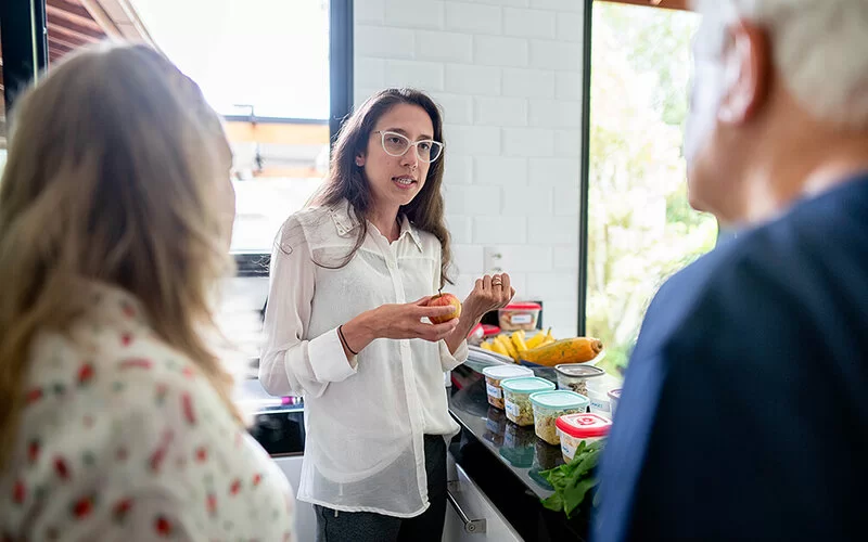 Eine Ernährungsberaterin steht in einer Küche und hält einen Apfel in der Hand. Sie berät ein älteres Paar, das nur von hinten und verschwommen zu sehen ist. Auf der Küchenzeile stehen eine Schüssel mit Bananen und verschiedene Plastikdosen mit Lebensmitteln.