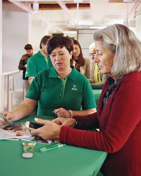 Eine AOK-Mitarbeiterin steht an einem Messetisch und spricht mit einer Interessentin