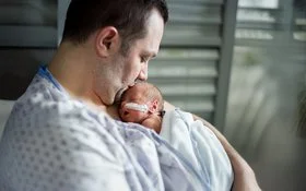 Vater mit nackter Brust hält schlafendes Frühchen mit nasaler Sonde eng an sich; sanfte Beleuchtung im Klinikzimmer.