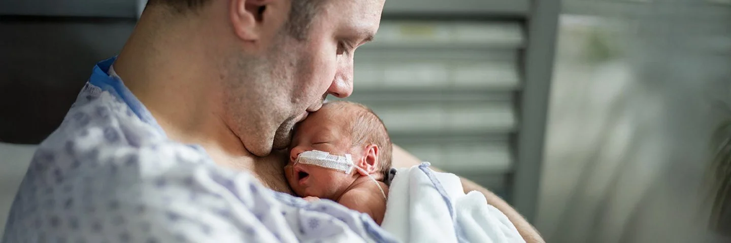 Vater mit nackter Brust hält schlafendes Frühchen mit nasaler Sonde eng an sich; sanfte Beleuchtung im Klinikzimmer.