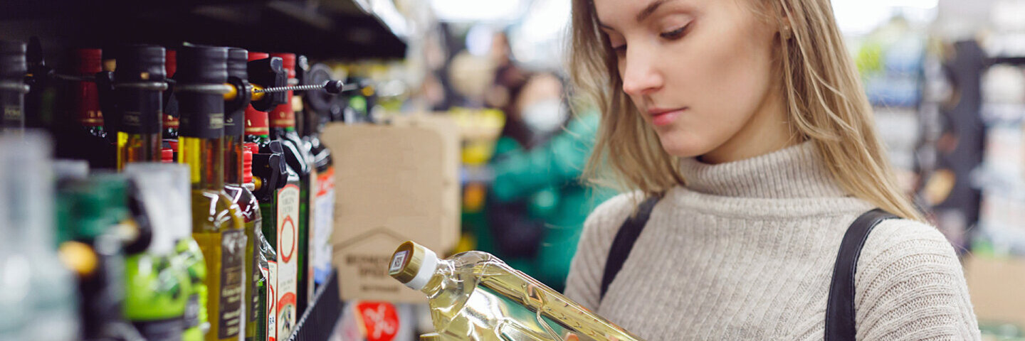 Eine junge Frau vergleicht im Supermarkt die Etiketten von zwei Ölflaschen.