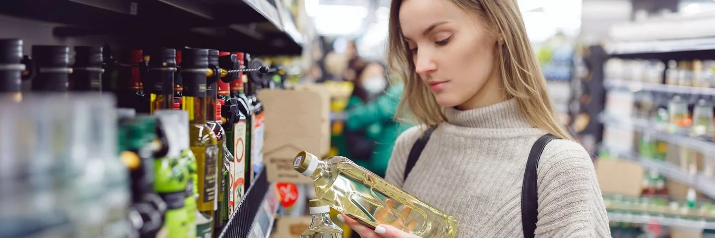 Eine junge Frau vergleicht im Supermarkt die Etiketten von zwei Ölflaschen.